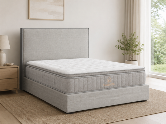 A light gray upholstered bed frame with a matching headboard and mattress, shown in a bright bedroom setting with a window, plant, and bedside table.